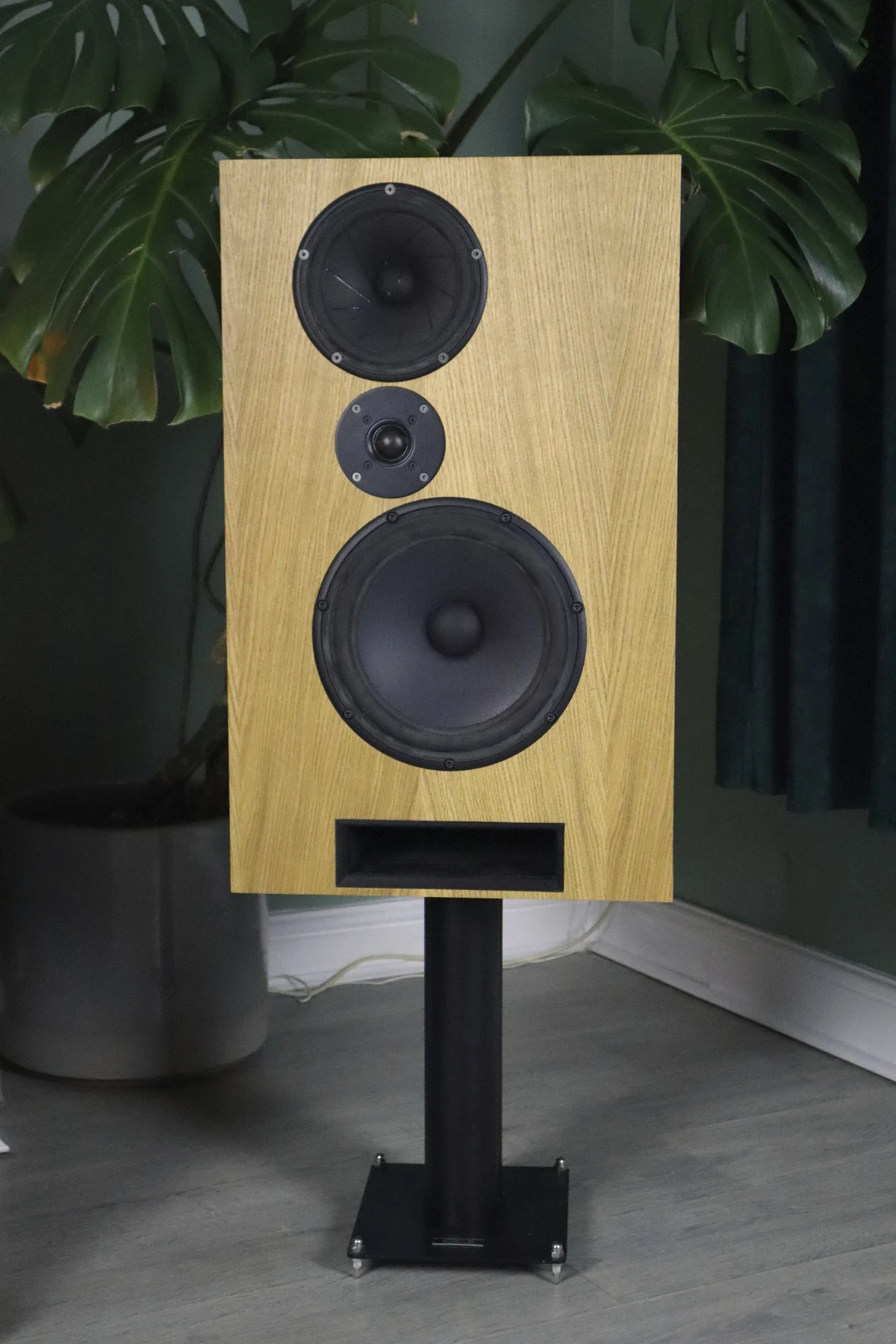 Front view of the MÆRE Cwen loudspeaker in natural oak on a black stand